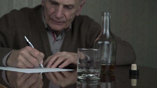 aged man drinks whiskey from transparent glass sitting at brown wooden table and writing testament on paper in dusky room