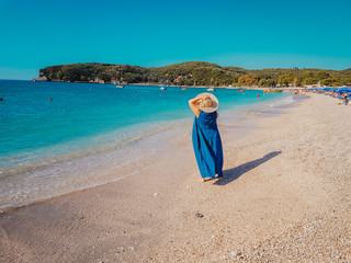 Fototapeta na wymiar Back view of the young woman in straw hat and blue dress relaxing on the beach and looking forward into the lagoon in Parga Greece.