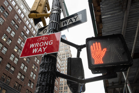 Signs on street corner that say Wrong Way, One Way and a red hand stop light.
