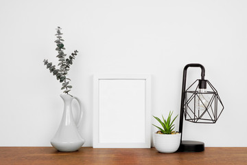 Mock up white portrait frame with home decor and potted plant. Wooden shelf against s a white wall. Copy space.