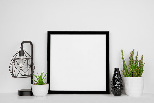 Mock up black square frame with home decor and potted plants. White shelf and wall. Copy space.