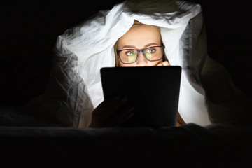 Woman in glasses lying in bed under the blanket using tablet late at night, can not sleep, addict...