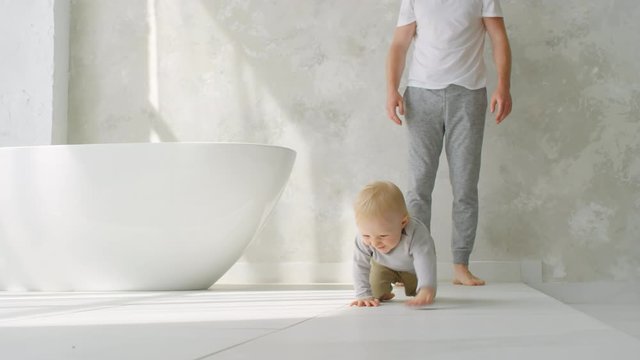 Ground-level shot of happy toddler girl crawling on bathroom floor towards camera and giggling. Unrecognizable father standing in background
