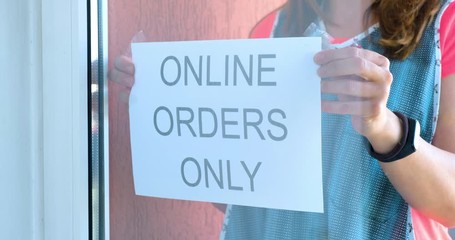Restaurant or shop owner wearing trendy apron, places 'Online Orders Only' sign in window, a common practice derived from safety measures during the 2020 Coronavirus pandemic.