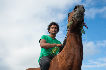 Young Maori man in green shirt riding brown horse along beach. - obrazy, fototapety, plakaty