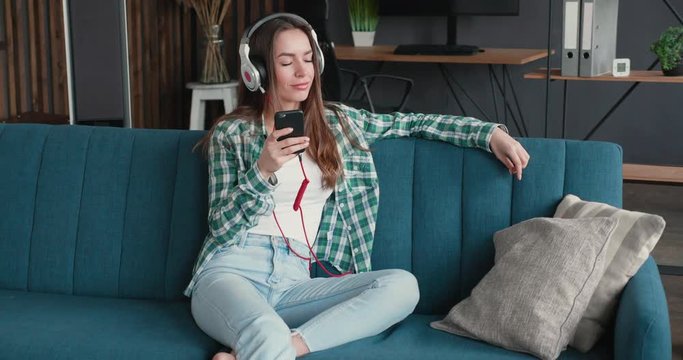 Attractive Woman is turning on her Favourite Music and started to relax on the Sofa. Young casual Woman is listening to Music in big Headphones, using Musically Apps for Smartphones. Music. Good Mood.