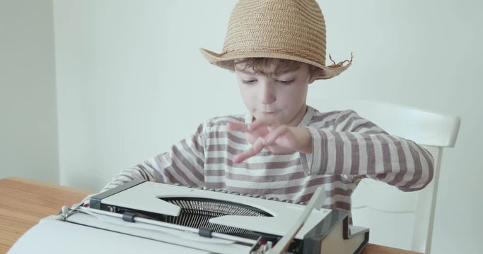 young boy counts as he types on an old vintage portable typewriter. He stops to check what he has typed before typing again with one finger. He is wearing a hat
