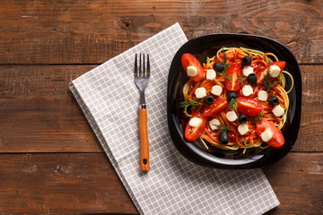 A black plate with spaghetti in a sauce decorated with cherry tomatoes olives and mozzarella on a linen napkin. Save a place.