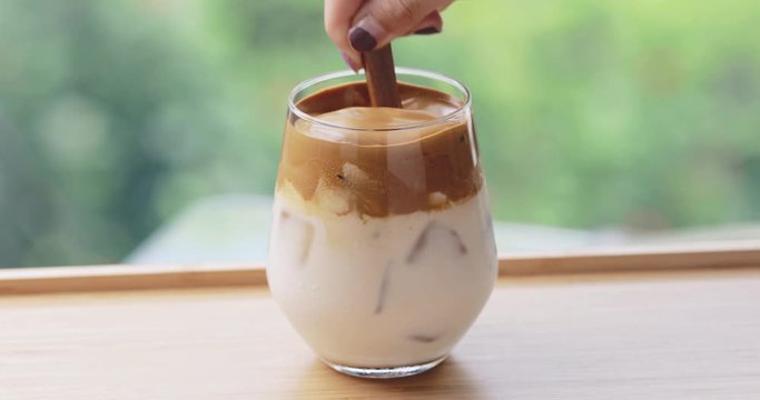 Dalgona Coffee Iced - Stiring and mixing fluffy creamy whipped coffee with milk and ice on wooden table in glass mug closeup in a cafe outdoor setting against trees background - Recipe video tutorial