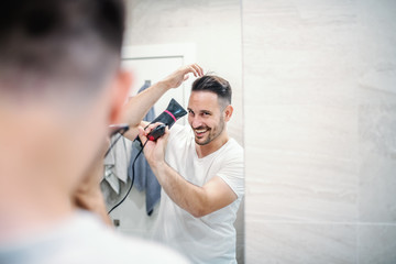 Attractive smiling caucasian unshaven man in pajamas standing in front of mirror and using hairdryer in bathroom.