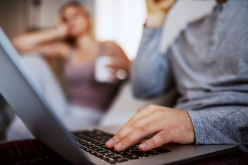 Close up of young man using laptop for internet surf. Woman in background sitting and getting bored because her boyfriend ignoring her.