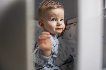 Portrait of charming blond little baby boy sitting in his crib and playing.