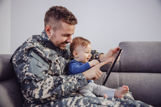 Smiling attractive bearded soldier sitting on sofa in living room with his little son and playing games on tablet.