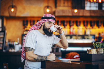 Young handsome tattooed bearded muslim hipster sitting in cafe in the morning and drinking his...