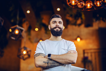 Low angle view of handsome muscular caucasian smiling tattooed bearded hipster standing in cafe with arms crossed.
