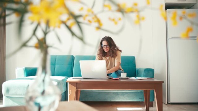 A young woman remote employee in glasses working from home with a laptop on a couch in a living room. Freelancer during self-isolation and quarantine