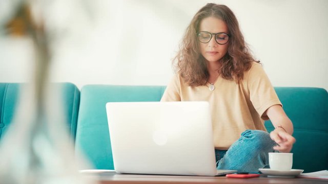 A young woman remote employee in glasses working from home with a laptop on a couch in a living room. Freelancer during self-isolation and quarantine