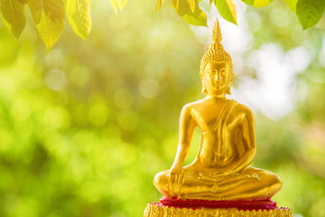 A golden Buddha statue sitting on a golden base with green blurred nature background with green leaves nature foreground, Buddhist Sabbath Day concept. - Powered by Adobe