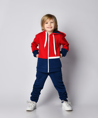 Happy smiling blond kid boy in red and blue sportwear and white sneakers poses holding hands at his waist