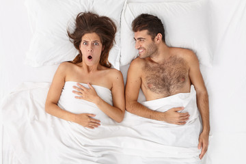 Obraz na płótnie Canvas Couple lying in bed under white blanket. Shocked woman overslept work, woke up very late, cheerful husband looks at surprised wife. Woman and man have lazy weekend after hard working week. Bed time
