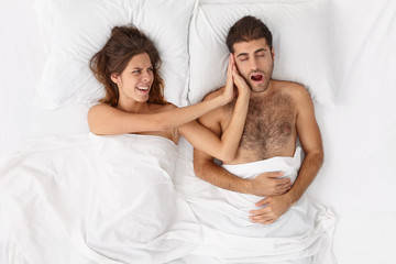 Frustrated annoyed woman cant stand mans snoring in bed during night, feels disturbed because of insomnia, stressed and irritated. Husband keeps wife awake with loud wheeze. Couple lifestyle