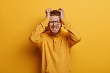 Tensed depressed man grabs head, feels despair, stands hopless indoor, wants to cry, expresses negative emotions, complains on bad life, unlucky fate, dressed in yellow clothes and spectacles