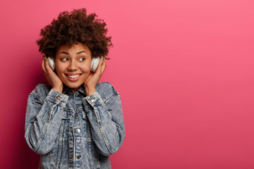 Delighted beautiful dark skinned woman listens music beat for energetic mood, smiles joyfully, looks aside, rests with joy, wears stereo wireless headphones, enjoys rthythm poses over rosy studio wall