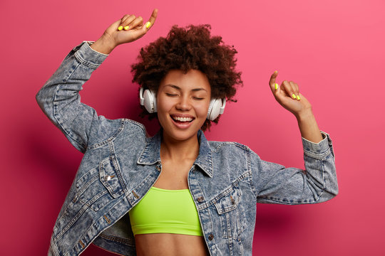 Joyful female dancer listens music in stereo headphones, enjoys every note, dances with raised arms, has happy dancing, dressed in top and fashionable denim jacket, smiles toothily, closes eyes