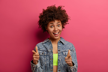 Cheerful curly adult woman picks you, makes great choice, greets someone with pleasant toothy smile, congratulates with good accomplishment, suggests join her team, poses indoor, gestures actively