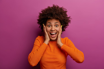 Happy energetic woman grabs face, hears wonderful news, realises she won huge money, stands impressed and speechless, dressed in orange jumper, feels lucky and thrilled, got incredible offer