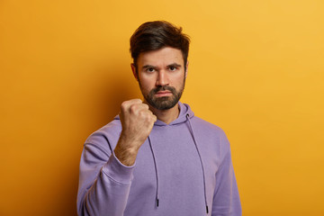 Annoyed aggressive Caucasian man shows fist, looses temper, looks at person with hate, promises to revenge, dressed in violet hoodie, isolated on yellow background. Negative emotions concept