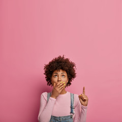Vertical shot of astounded woman covers mouth, stands speechless, shows something disturbing,...