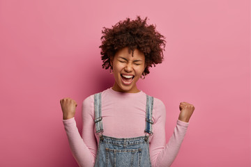 Cheerful lucky dark skinned model happy to achieve goal, clenches fists and exclaims with joy, becomes real champion, cheers over something, stands glad against pink background. Celebration.