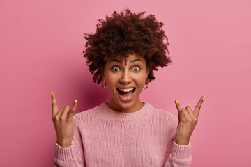 Fun and amusement. Joyful dark skinned young woman shows heavy metal gesture, foolishes around at concert, exclaims gladfully as hears favorite rock song, enjoys good loud sound, stands indoor