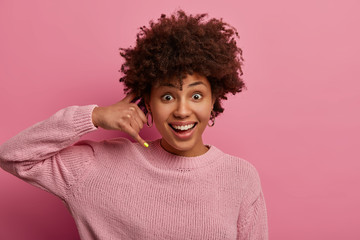 Give me call sometime. Pleased ethnic woman with Afro hair makes phone gesture, asks person for telephone number to be in contact, smiles positively, suggests you to hang, wears rosy jumper.