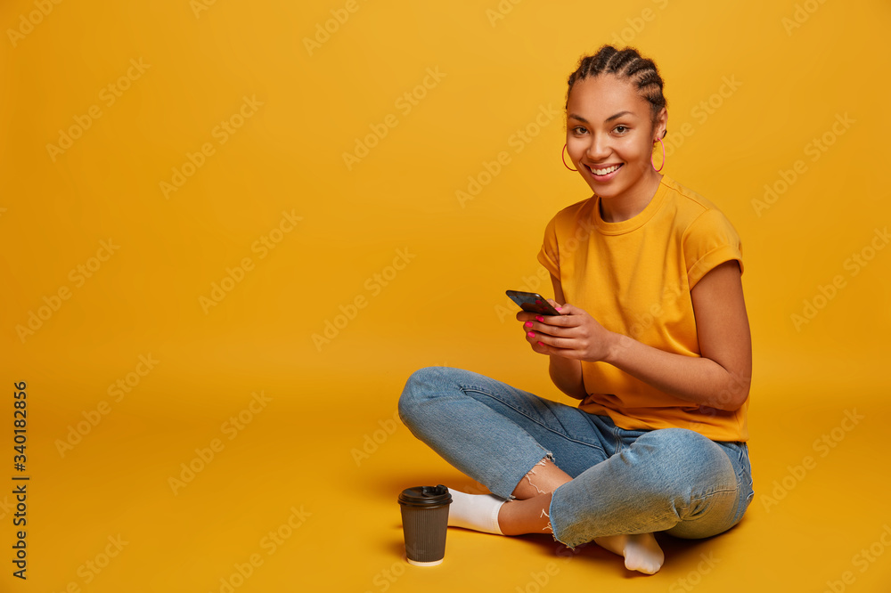Wall mural Photo of relaxed dark skinned girl happy to receive message from lover, holds mobile phone, watches funny cute video, sits in lotus pose, drinks takeaway coffee, isolated on yellow background - Wall murals