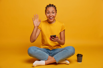 Happy dark skinned girl waves palm in hi, sits crossed legs, holds modern smartphone, uses application for texting with friends, stays in touch online, drinks coffee to go, isolated on yellow wall