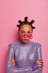 Indoor shot of serious Afro American female model stands with arms folded, looks calmly at camera, wears pink sunglasses, purple glittering jumper, confident in herself, isolated on pink backgroud