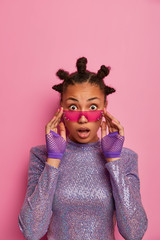 Astounded dark skinned hipster girl reacts on shocking relevation, gazes surprisingly through trendy rosy sunglasses, has hair buns, stands impressed indoor against pink pastel studio background