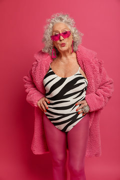 Vertical shot of mature woman with curly hair, keeps lips rounded, wants to kiss someone, wears fashionable outfit, looks through pink sunglasses, stands indoor. Retired lady relaxes at home