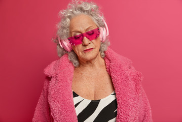 Portrait of serious thoughtful elderly woman wears massive headphones, listens melancholic music,...