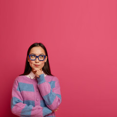 Indecisive pretty schoolgirl holds chin and looks up thoughtful, makes choice with serious expression, imagines plan, thinks about something important, wears big transparent glasses, warm jumper