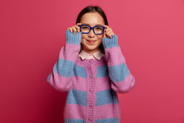 Portrait of pleasant looking female student looks through optical glasses, checks out something nice that captures attention, has interesting gaze, stands pleased, wears striped jumper, poses indoor