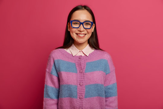 Half length shot of beautiful Asian schoolgirl wears big transparent glasses with blue frame, smiles with satisfaction and interest, happy to get excellent mark, isolated on pink bright background
