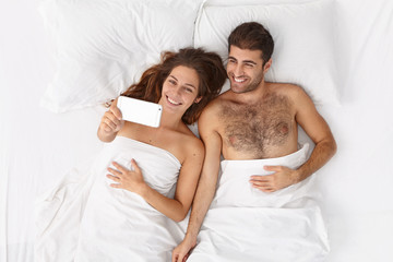 Obraz na płótnie Canvas Happy family couple make morning selfie, pose at camera of modern smartphone in bed, have fun and smile positively, woke up in good mood, use new cool online application, enjoy weekend at home