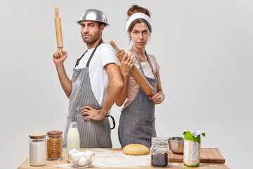 Serious woman and man stand back to each other, hold wooden rolling pins, participate in culinary...