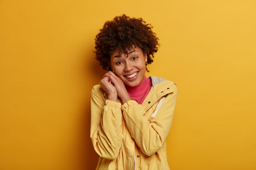 Fototapeta na wymiar Happy positive Afro American woman smiles gently, leans head on hands, glad to hear compliment, dressed in anorak, observes something pleasant, stays on bright side expresses enthusiasm and positivity