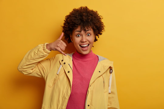Call me back. Friendly positive woman with curly hair, gestures near ear, shows phone gesture, looks cheerfully at camera, tries be in contact with friends or relatives, stands indoor over yellow wall