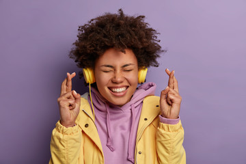 Wishful happy woman with natural curly hair anticipates relish and good news, crosses fingers and smiles broadly, awaits for dream come true, wears stereo headphones, listens pleasant music.