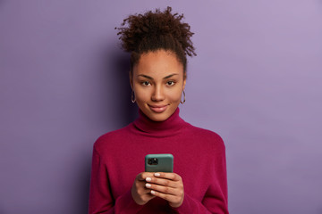 Photo of pleased dark skinned millennial girl holds mobile phone, waits for call, texts with friend in online chat, uses special application, taps smartphone screen, checks newsfeed, browses internet